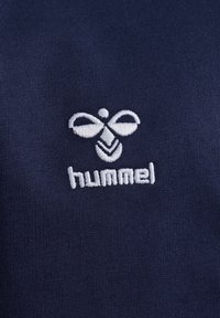 Navyblaue Stoff mit einer glatten Textur, versehen mit einem weißen gestickten Logo im bienenähnlichen Design und dem Wort "hummel" darunter.