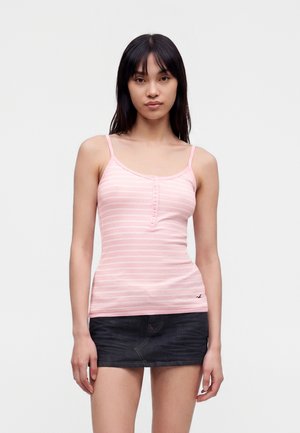 Junge Frau mit glatten schwarzen Haaren, die ein pink gestreiftes Tanktop und einen dunklen Jeans-Minirock trägt, vor einem schlichten Hintergrund stehend.