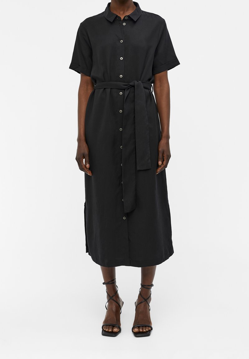 Femme portant une robe midi noire à boutons, manches courtes et ceinture nouée à la taille, associée à des sandales noires à talons hauts et fines lanières.