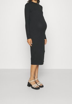 Kobieta w ciąży ubrana w dopasowaną czarną sukienkę midi z długim rękawem oraz białe sandały na słupku z paskami, stojąca na jednolitym tle.