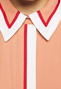 Camicia color pesca con colletto bianco bordato di rosso e striscia verticale rossa. Tessuto liscio, design classico e forma del colletto ben definita.