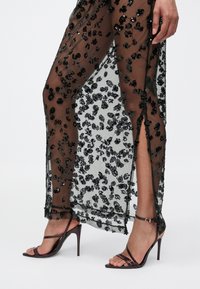 Benen in een doorschijnende zwarte jurk met bloemenpailletten en een split aan de zijkant, gedragen met donkerbruine sandalen met hoge hakken en bandjes, op een witte achtergrond.