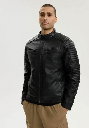 Mężczyzna w czarnej skórzanej kurtce na zamek z pikowanymi detalami na ramionach i rękawach, ubrany w jasnobrązowe spodnie, stojący na neutralnym tle.