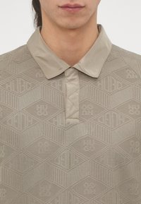 Beige gallér ing, amely hexagonokból és vonalakból álló texturált mintával rendelkezik, eltérő sima gallérral és gombolású résszel.