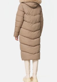 Długa beżowa puchowa kurtka z poziomym przeszyciem w kształcie jodełki, kapturem wyłożonym futrem i dopasowaną sylwetką, zestawiona z białymi kozakami sięgającymi kolan.