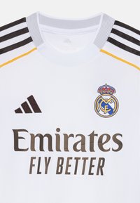 Balts sporta krekls ar apaļu kakla izgriezumu, kam ir melnas svītras uz pleciem, zila un zelta izšūta.logo un brūna teksta.