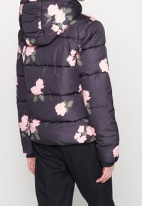 Prošívaná bunda tmavě fialové barvy s velkými růžovými květinovými vzory. Má kapuci a prošitý design s hladkým saténovým vzhledem.