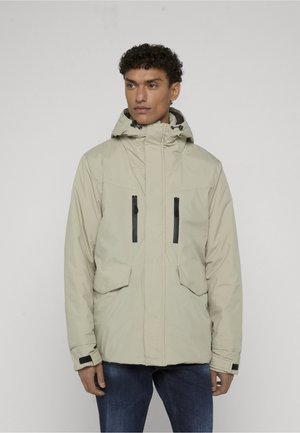 Homme portant une veste d'hiver beige à capuche avec des poches poitrine zippées et des poignets ajustables, debout devant un fond uni.