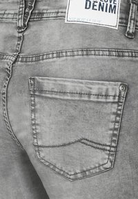 Pantalones de mezclilla gris con un bolsillo trasero único con un contorno cosido, detalle de botón de metal y una etiqueta de tela en la cinturilla.