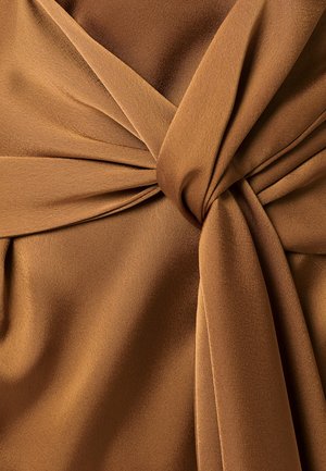 Gladek rjav tkanina, tesno zavit v vozel, ustvarja mehke gube in teksturne variacije v topli osvetlitvi.