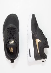 Black athletic shoes with a mesh upper, gold Nike swoosh accent, white sole, and visible air cushioning. Features laces and pull tab.
