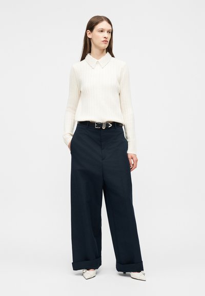 Femme debout portant un pull blanc avec un col orné de perles, un pantalon large noir, une ceinture noire et des chaussures blanches à bout pointu.