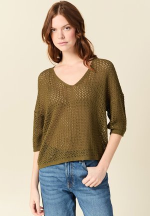 Giovane donna con capelli castano ramati mossi, che indossa un maglione a V a maglia aperta verde oliva e jeans blu, in piedi davanti a uno sfondo chiaro.