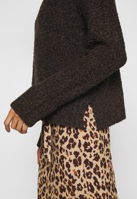 Brauner gestrickter Pullover mit strukturiertem Finish und seitlichem Schlitz. Er wird über einem beigen Rock mit Leoprint getragen.