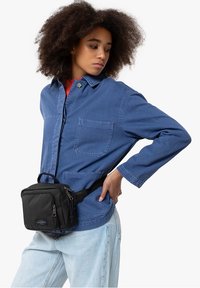 Giovane con i capelli ricci che indossa una giacca blu, jeans chiari e una borsa a vita nera, in posa con una mano sulla vita.