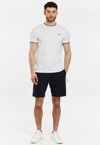 Camiseta blanca de cuello redondo con ribete azul marino, combinada con pantalones cortos azul marino. El modelo lleva zapatillas blancas. Diseño simple, material de algodón, aspecto casual.