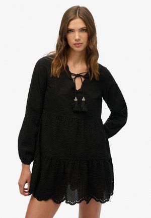 Vestido corto negro con tejido texturizado, cuello redondo con detalle de lazo, mangas largas y un fondo acampanado con dobladillo ondulado.