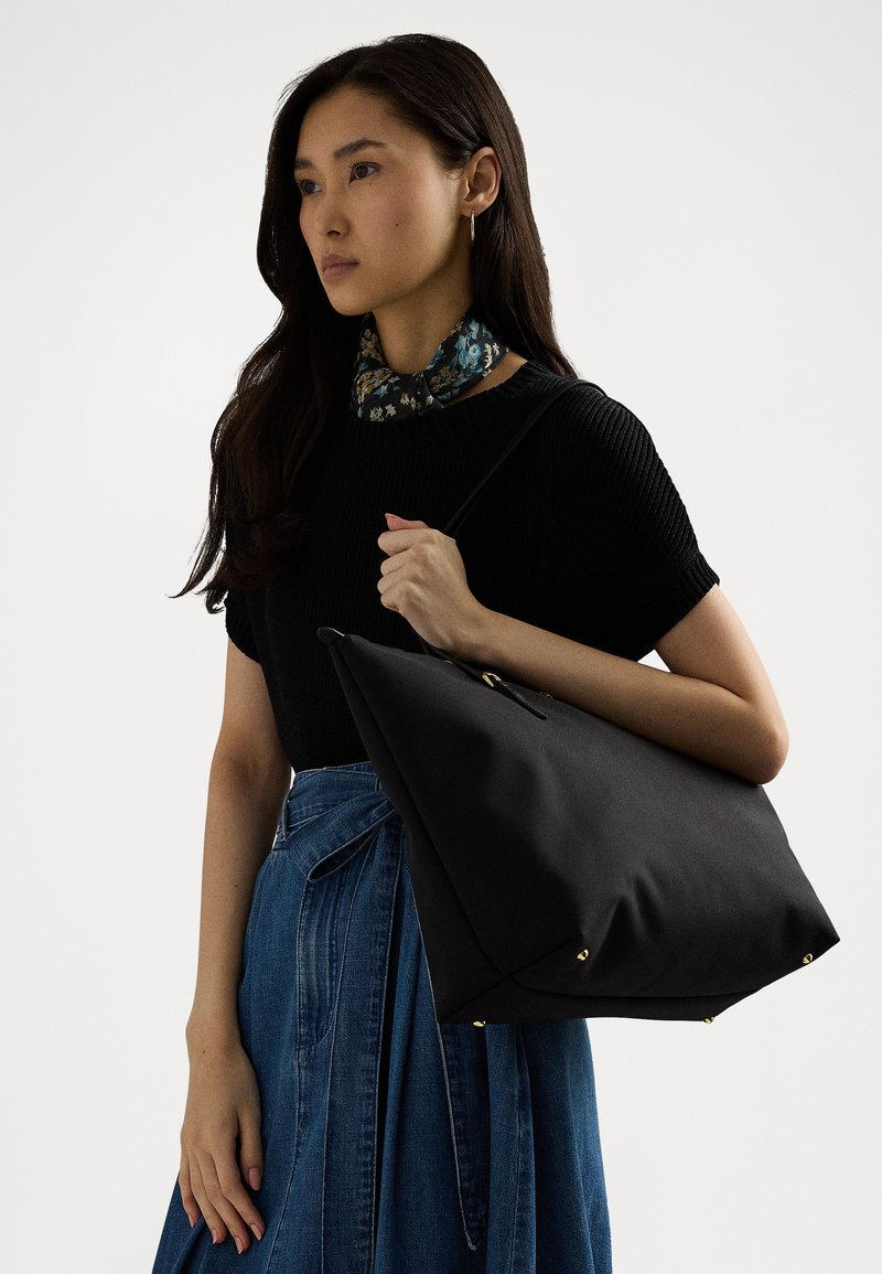 Svart tote bag med en strukturert form, gullfarget hardware-accents og en teksturert overflate. Bært på skulderen av en person i uformell antrekk.