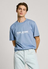 Light blue t-shirt with white "PEPE JEANS" graphic, round neck, short sleeves, paired with light denim jeans. Smooth cotton texture.