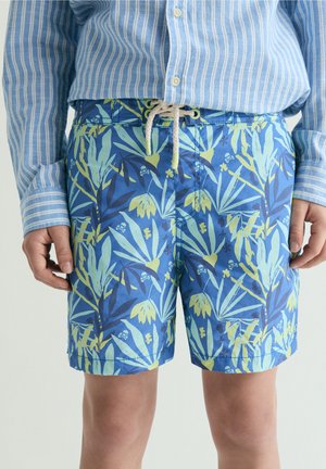 Pantalones cortos con estampado tropical azul con calaveras y hojas verde claro, usados con una camisa abotonada de rayas azules y blancas.