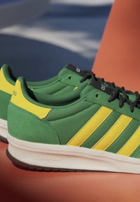 Green and yellow Adidas sneakers with black laces resting on an orange surface, featuring Cloudfoam cushioned white midsoles.