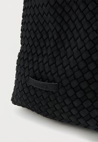 Μαύρη υφασμένη τσάντα με υφή στην επιφάνεια, που διαθέτει μια λεία μαύρη ετικέτα προσαρτημένη. Ο σχεδιασμός έχει δομημένο, στρογγυλό σχήμα.