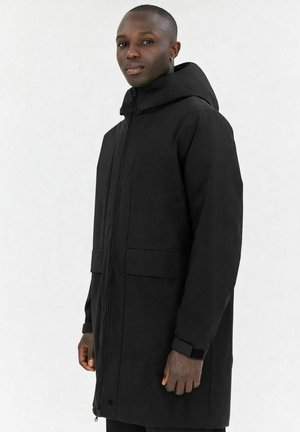 Mand iført en sort vinterjakke med hætte og forlommer, stående mod en ensfarvet hvid baggrund, vendt let til siden.