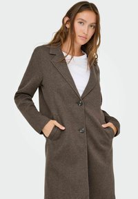 Bruine, lange jas met een op maat gemaakte uitstraling, twee voorzakken, zwarte knopen en een gestructureerde stof. Draag over een wit shirt.