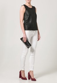 Czarny skórzany top bez rękawów zestawiony z białymi dopasowanymi jeansami i bordowymi butami na wysokim obcasie, trzymający czarną kopertówkę z kontrastującym akcentem.