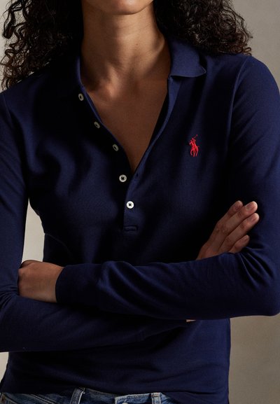 Tamsiai mėlynas polo marškinėliai ilgomis rankovėmis iš tekstūruotos medžiagos, su trimis mygtukais ir mažu raudonu išsiuvinėtu logotipu kairėje krūtinės pusėje.