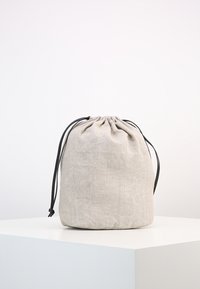 Beige linnen trekkoordzak met zwarte koorden, geplaatst op een wit voetstuk tegen een eenvoudige lichte achtergrond.