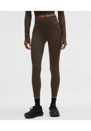 Persoon draagt hoge bruine leggings en een crop top met lange mouwen, gecombineerd met zwarte sportschoenen en sokken, staand tegen een effen achtergrond.