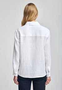 Chemise en lin blanc avec un col, des manches longues et des poignets boutonnés. Le tissu a une légère texture, lui donnant un aspect décontracté.