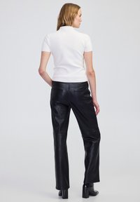 Eine Frau, die mit dem Rücken zur Kamera steht und ein weißes kurzärmeliges Oberteil, schwarze Lederhosen und schwarze Absatzstiefeletten trägt, vor einem einfarbigen Hintergrund.