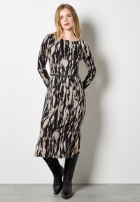 Langarmkleid mit einem schwarz-beigen abstrakten Muster, tailliertem Schnitt, seitlichen Schlitzen und kombiniert mit schwarzen kniehohen Stiefeln.