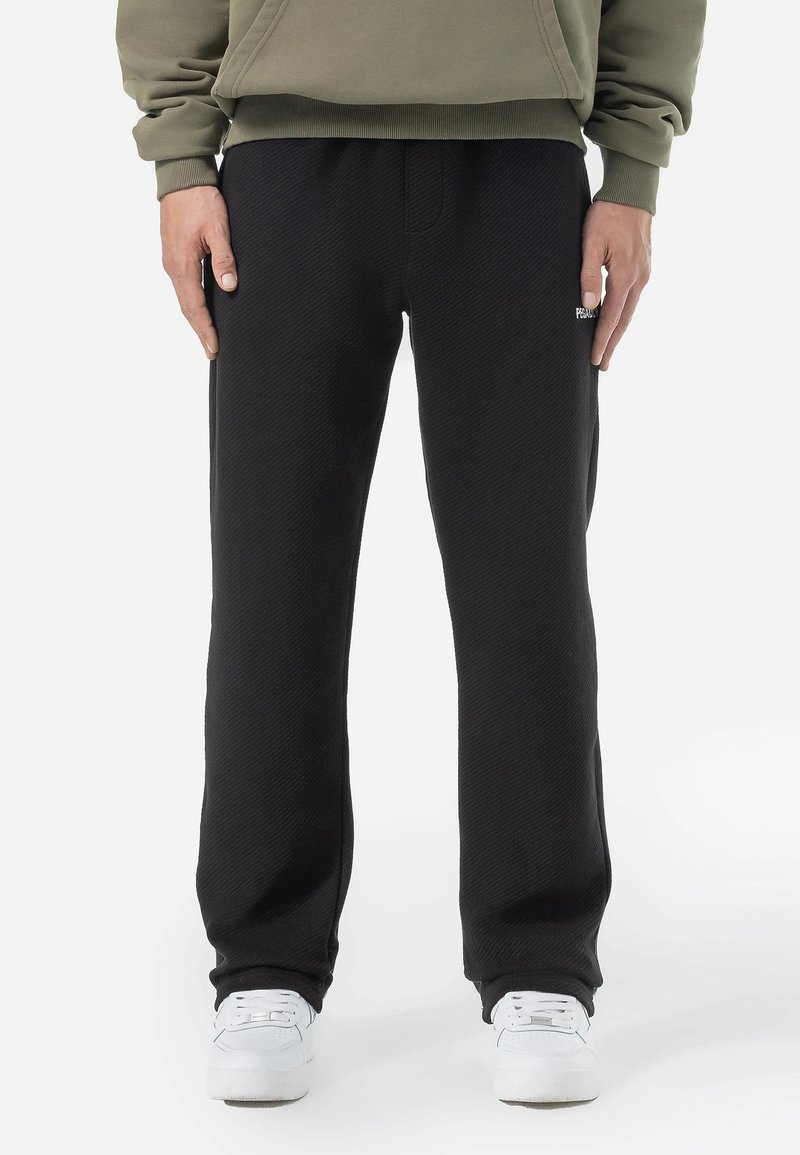Pantalones de chándal negros con textura y ajuste suelto, que cuentan con bolsillos laterales y una cintura elástica. Llevados con zapatillas blancas.