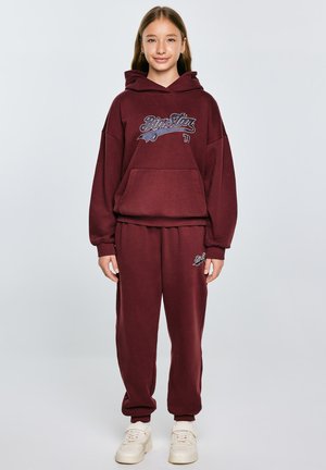 Bordowa bluza z dużą kieszenią z przodu i graficznym napisem, zestawiona z pasującymi spodniami dresowymi z detalem logo. Białe sneakersy dopełniają stylizację.