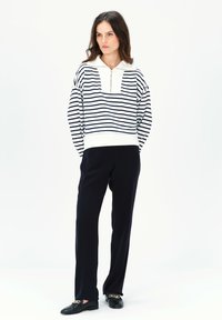 Pullover bianco e blu a righe con colletto con zip, abbinato a pantaloni neri e scarpe basse con accentuazione in catena dorata.