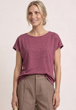 Uśmiechnięta kobieta o blond włosach związanych do tyłu, ubrana w bordowy krótki rękaw włożony do beżowych spodni z wysokim stanem, trzyma plecioną kopertówkę.