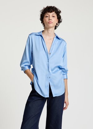 Hellblaue Satinbluse mit langen Ärmeln, Knopfleiste vorne, spitzem Kragen und lockerem Schnitt, kombiniert mit dunklen Jeanshosen.