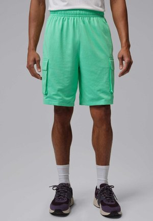 Man met mintgroene cargoshorts, witte sokken en donkerpaarse hardloopschoenen staat tegen een effen grijze achtergrond.