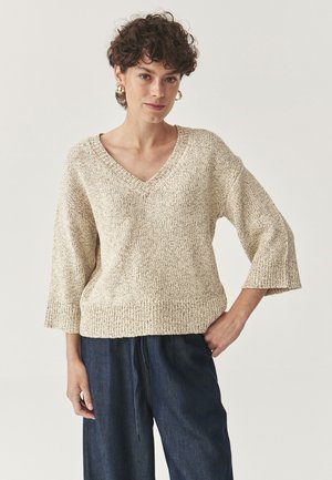 Beige V-hals sweater med tekstureret strik, brede ærmer og en ribbet kant, parret med mørke denimbukser. Enkel og afslappet design.