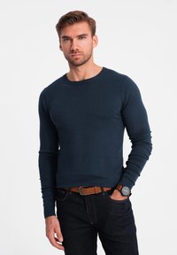 Mörkblå stickad tröja med rund halsringning och långa ärmar, med en slim fit. Kombinerad med mörka jeans och ett brunt bälte.