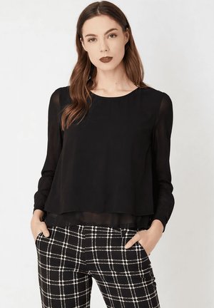 Mujer con cabello largo y castaño que lleva una blusa negra transparente de manga larga y pantalones a cuadros en blanco y negro, manos en los bolsillos.