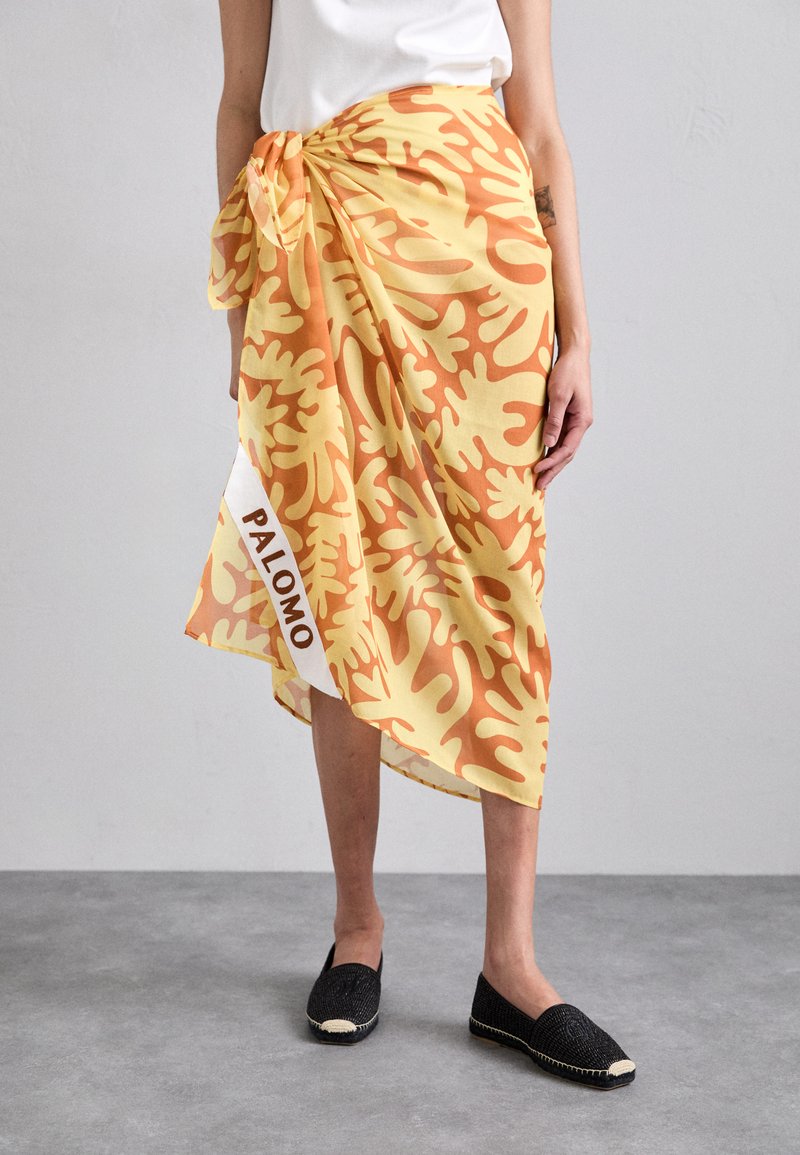 Wrap skirt in yellow and orange, featuring a bold leaf pattern, tied at the waist, with a side panel displaying "PALOMO" in white.