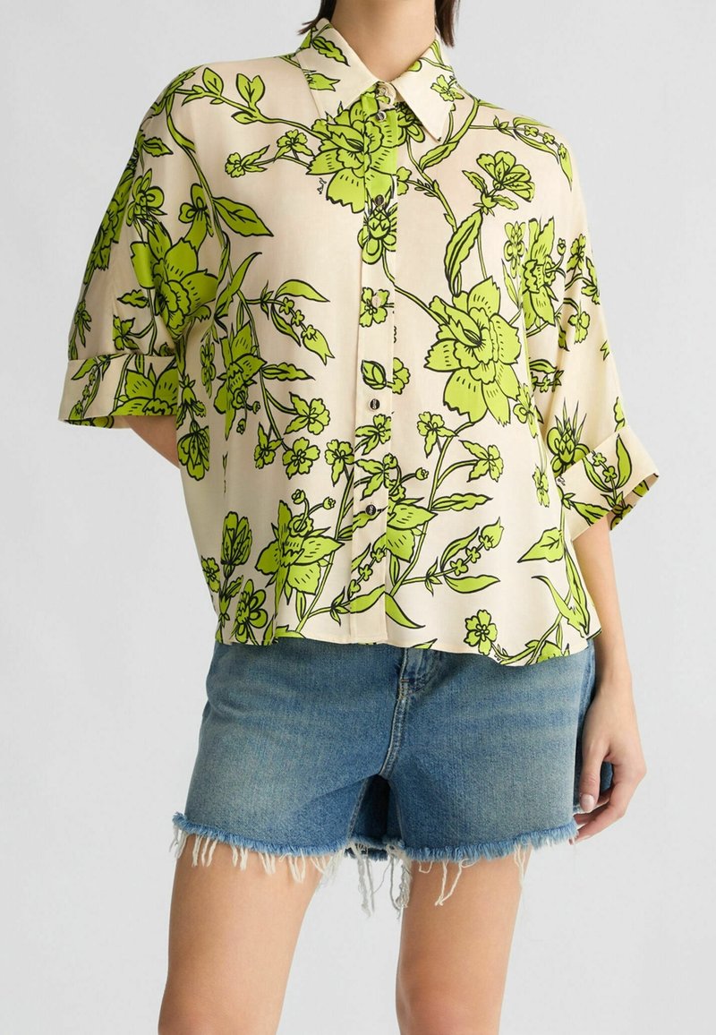 Femme portant une chemise beige à boutons avec un grand imprimé floral vert et des manches retroussées, associée à un short en jean effiloché.