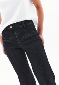 Mustat farkut, joissa on korkea vyötärö, etutaskut, näkyvät ompeleet ja hopeinen napin kiinnitys. Silkkinen rakenne, suora lahje.