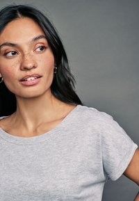 T-shirt gris à manches courtes avec un col décontracté, présenté avec une texture de tissu douce. Le modèle présente des taches de rousseur visibles et des boucles d'oreilles créoles.