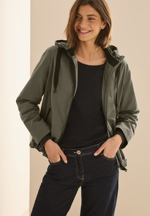 Giacca verde oliva con cappuccio, chiusura a cerniera, polsini elasticizzati e orlo regolabile. Indossata sopra una maglia nera, abbinata a jeans scuri.