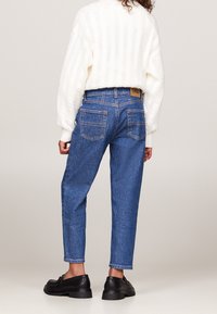 Jeans de mezclilla azul con un corte relajado, que cuentan con una cintura alta, cinco bolsillos y un parche de cuero beige en la parte trasera. Combinados con zapatos negros.